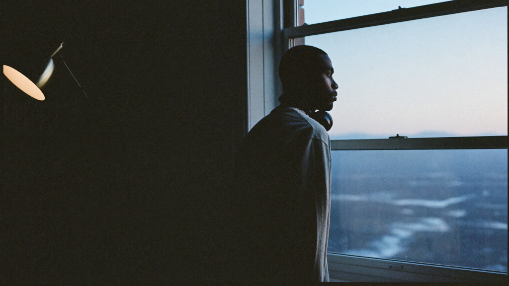 Independent artist standing at a home studio window at dawn with headphones around his neck
