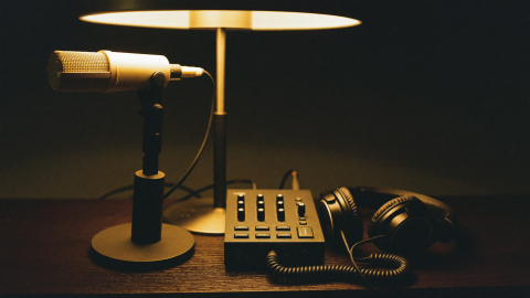 A microphone on a stand, a desktop audio interface and studio headphones on a dark desk under a warm lamp