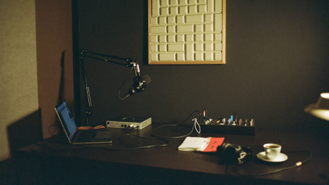 A lived-in home studio desk with a laptop, audio interface, microphone on a boom arm, headphones and a coffee cup