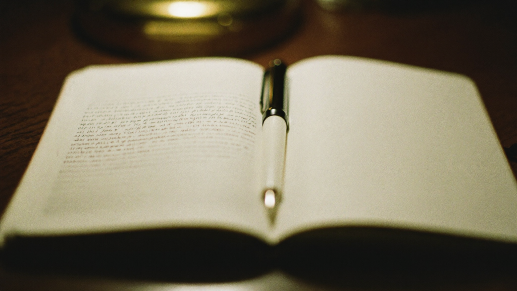 A pen resting across an open notebook on a dark desk under a warm lamp