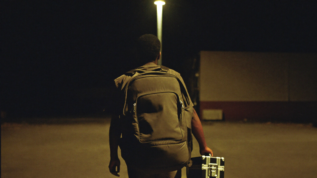 Independent artist carrying a gear bag and hard case through a dim parking area at night