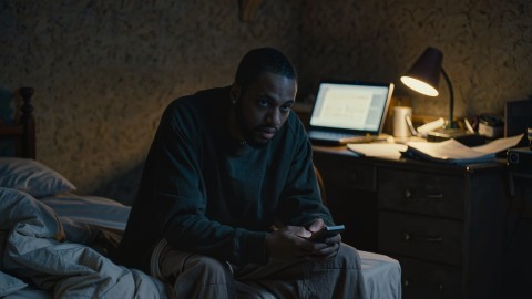 Why I Stopped Chasing Playlists (And What I Do Instead) 13 Independent artist sitting on the edge of his bed at night holding a phone, laptop glowing on the desk behind him